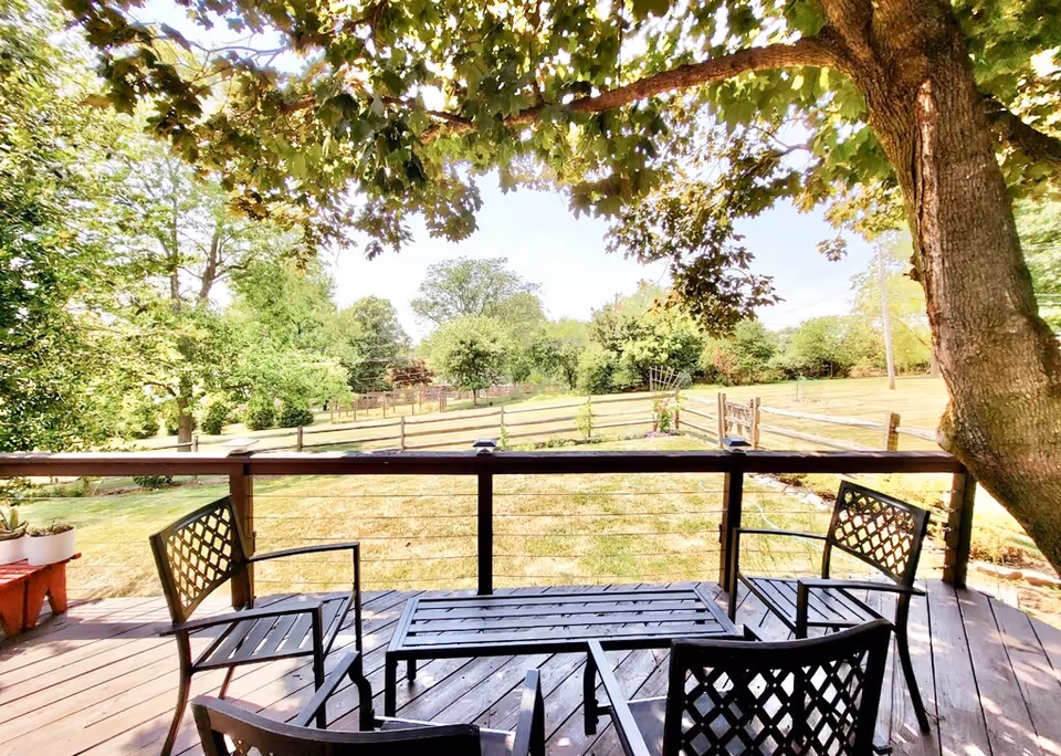 A wooden deck with four black metal chairs and a matching table, shaded by a large tree. Beyond the deck is a fenced grassy yard with various trees and shrubs under a clear sky.
