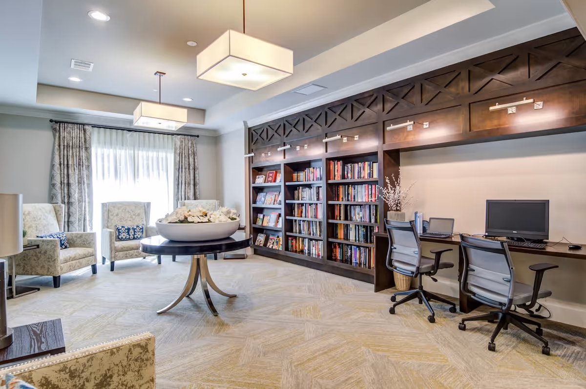 A bright and spacious room with large windows covered by sheer curtains, two upholstered armchairs with patterned cushions, a round table with a decorative bowl of flowers in the center, a built-in dark wood bookshelf filled with books, and a desk area with two office chairs and computer monitors.