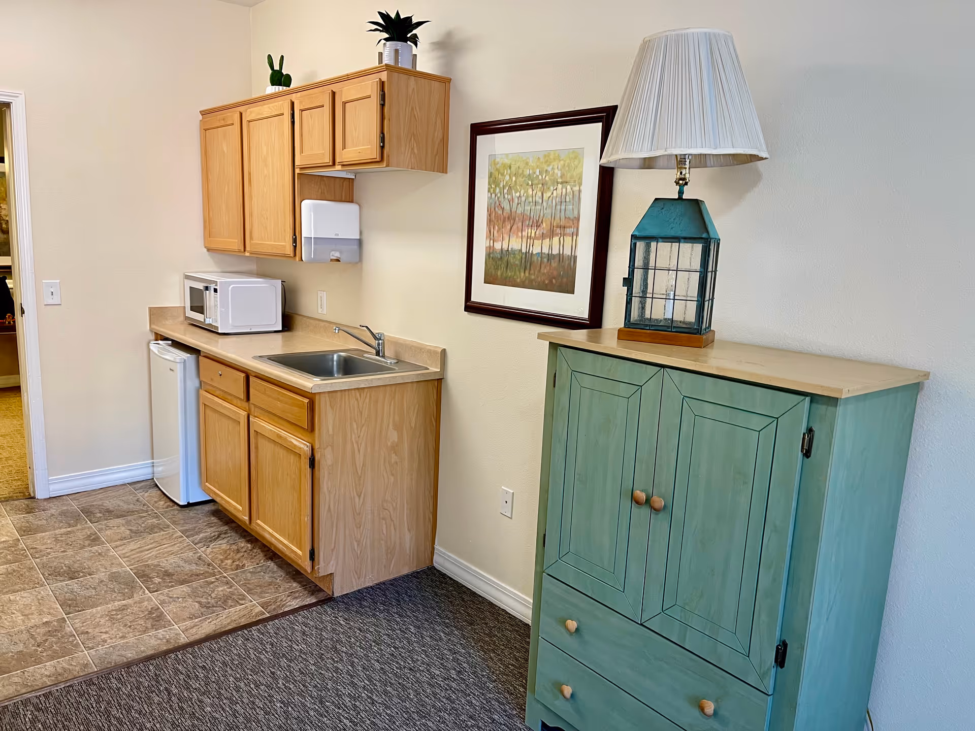 Small kitchenette with wooden cabinets, a sink, microwave and mini fridge next to a green storage cabinet topped with a lamp and framed artwork.