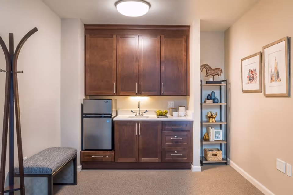 A small kitchenette area with dark wooden cabinets, a mini refrigerator, a sink, and a countertop with cups and a bowl of green apples. To the left, there is a coat rack and a cushioned bench. On the right side, there is a shelving unit with decorative items and two framed pictures hanging on the wall.