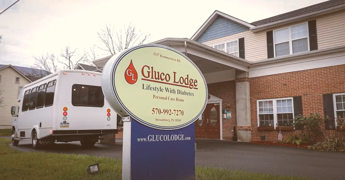 Exterior view of Gluco Lodge Personal Care Home building with a large sign in front displaying the facility name, slogan 'Lifestyle With Diabetes', contact phone number, and website. A white shuttle bus is parked near the entrance.