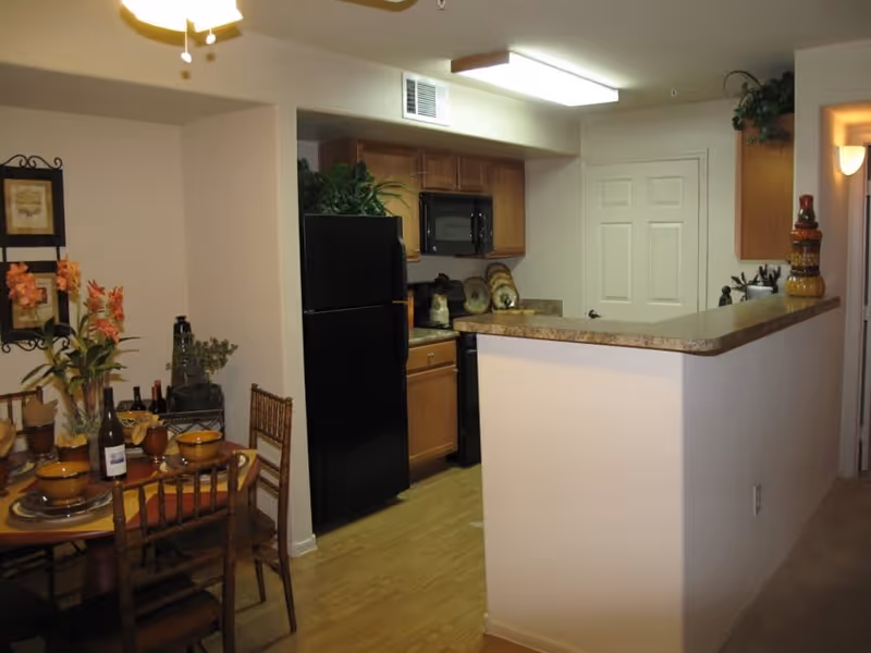 Interior view of a senior living facility showing a kitchen area with wooden cabinets, a black refrigerator, a microwave, and a countertop bar. Adjacent to the kitchen is a dining area with a round table set with dishes, bowls, wine bottles, and floral decorations. The room has light-colored walls and wooden flooring.