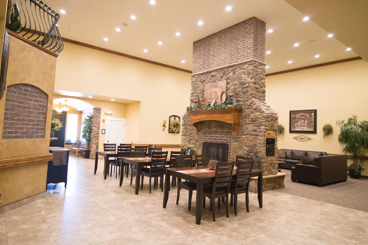 Spacious assisted-living common area with multiple dining tables and chairs arranged around a large stone fireplace and adjoining lounge seating.