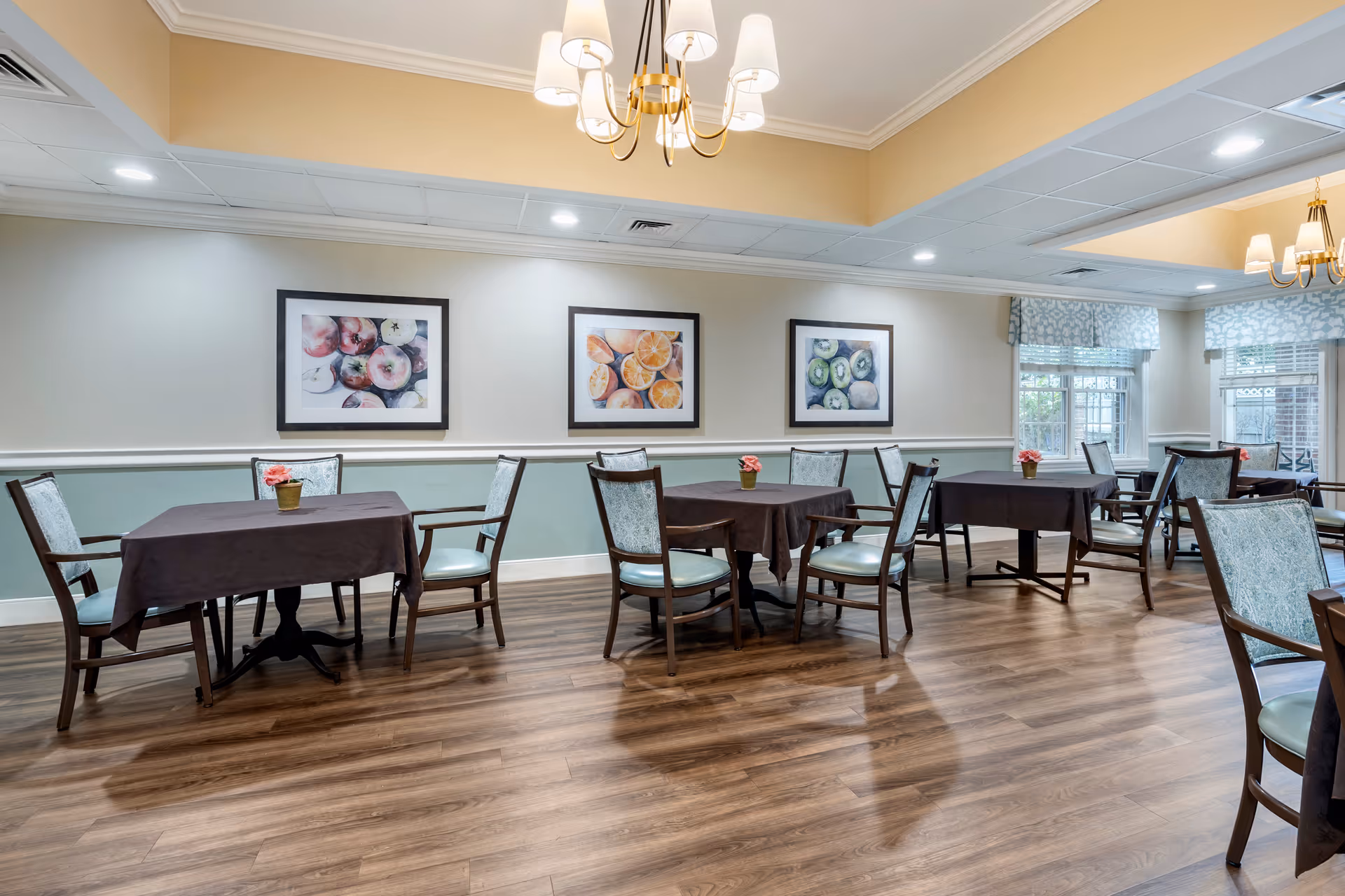 A dining room with several tables covered in dark tablecloths, each surrounded by wooden chairs with light blue patterned cushions. The room has wood flooring, light-colored walls with a chair rail, and three framed pictures of fruit on the wall. There are windows with patterned valances letting in natural light and ceiling lights including chandeliers.