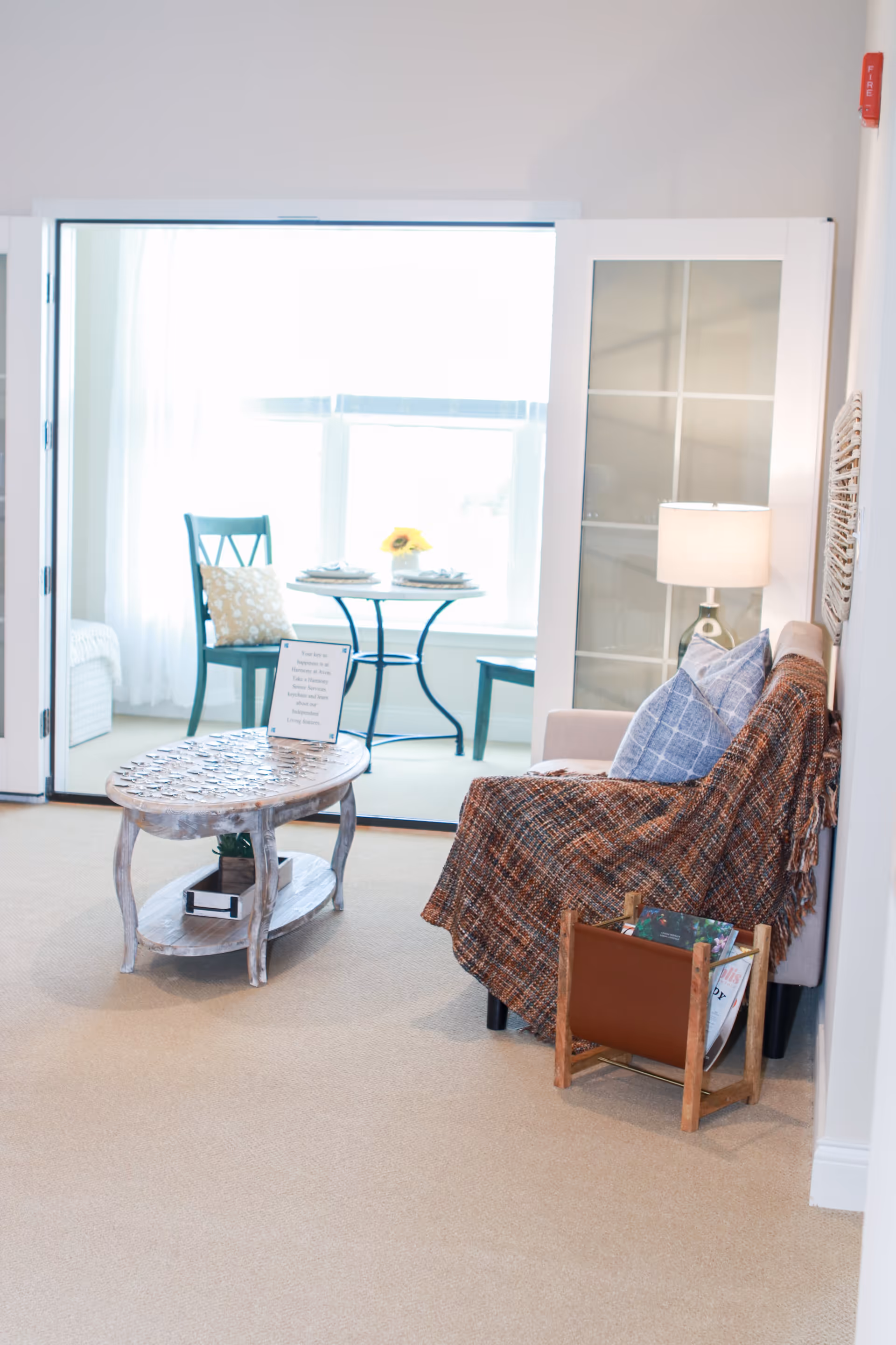 A cozy living room area with a beige couch covered by a brown and orange plaid throw blanket and two blue patterned pillows. Next to the couch is a wooden magazine rack holding magazines. A small wooden coffee table with a decorative top and a sign on it is in front of the couch. In the background, there is a small dining area with a round table set for two, two green chairs, and a window letting in bright natural light. A lamp with a beige shade is on a side table next to the couch.
