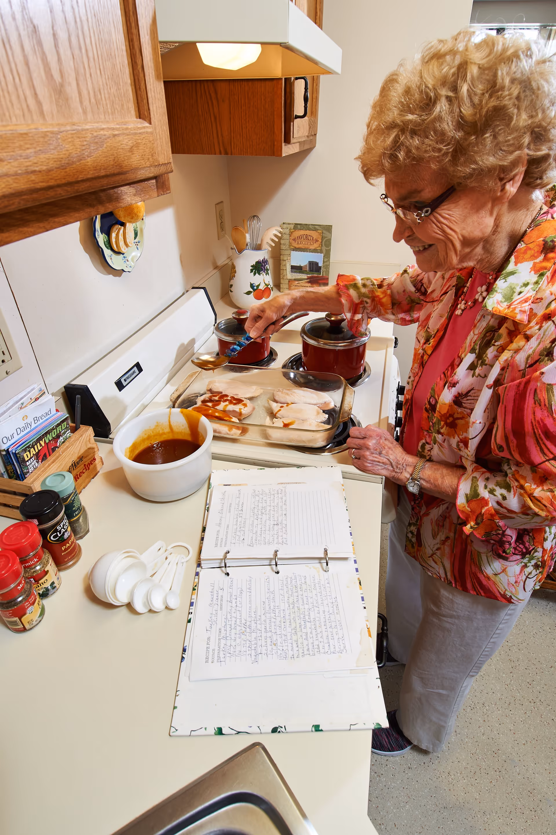 An elderly woman with curly hair and glasses is standing in a kitchen, spooning sauce onto chicken breasts in a glass baking dish on the stove. The kitchen counter has various spices, measuring spoons, a bowl of sauce, and an open recipe binder. The woman is smiling and wearing a floral patterned jacket over a pink top.