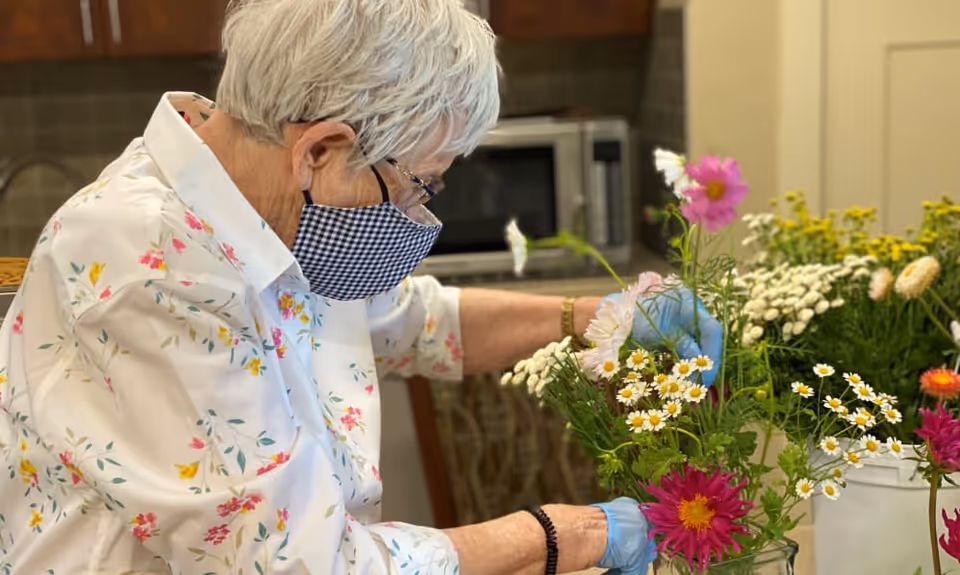 An elderly woman wearing a floral shirt, a checkered face mask, and blue gloves is arranging colorful flowers in a vase inside a room with a microwave in the background.