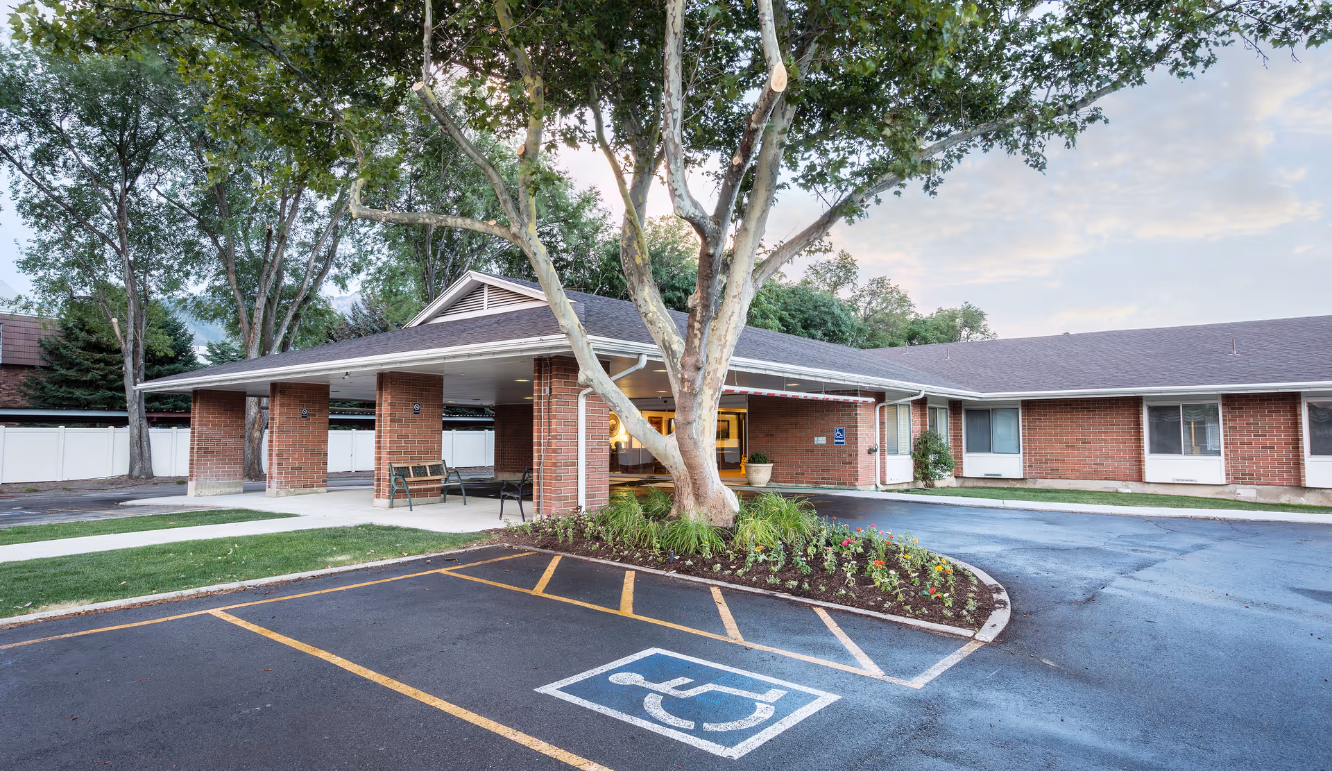 Single-story brick building entrance with a covered portico, accessible parking space, large tree, and landscaped flowerbed.