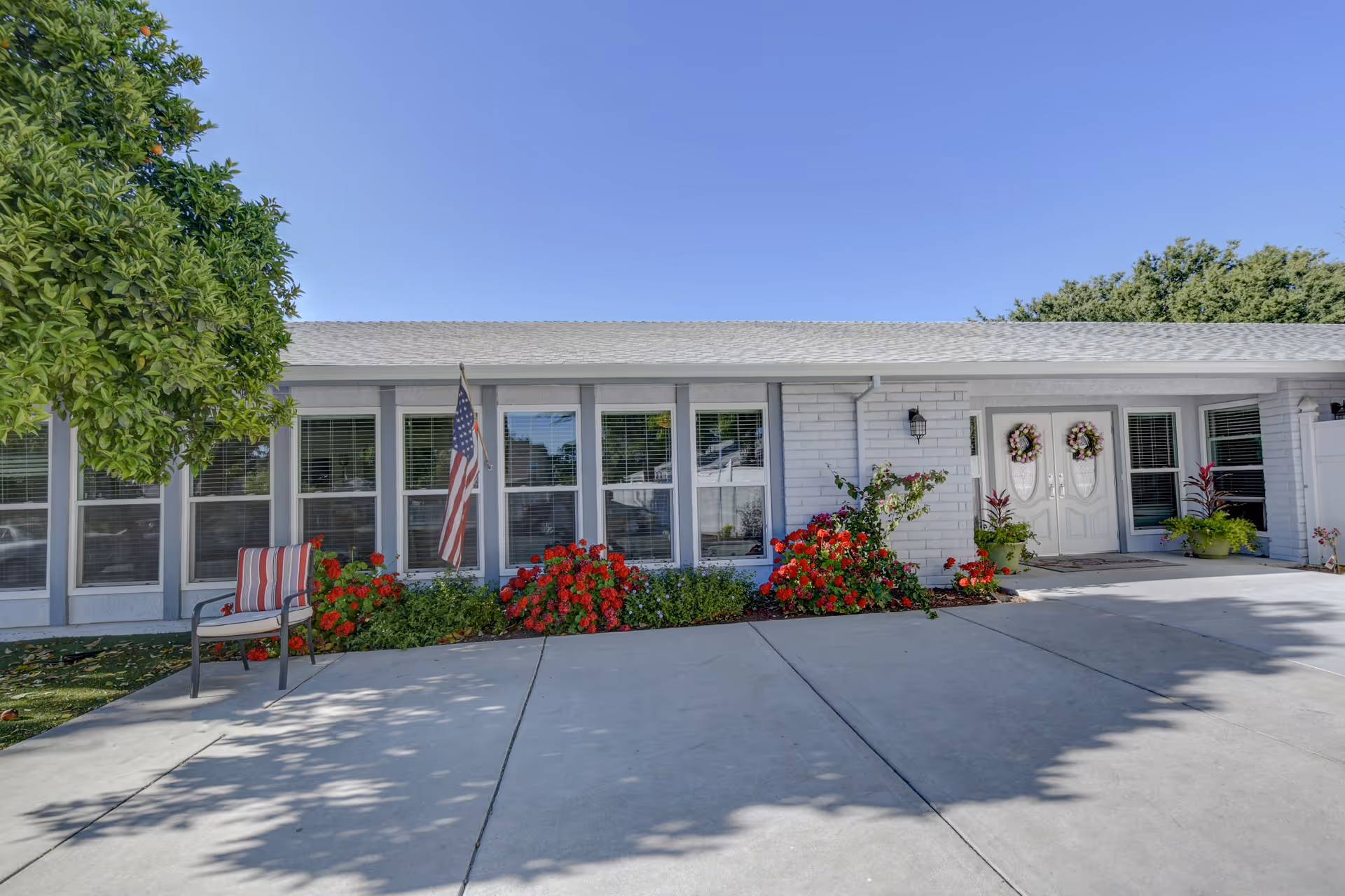 Front of a single-story assisted living building with double doors, an American flag, red flowering bushes, and a chair by the driveway.