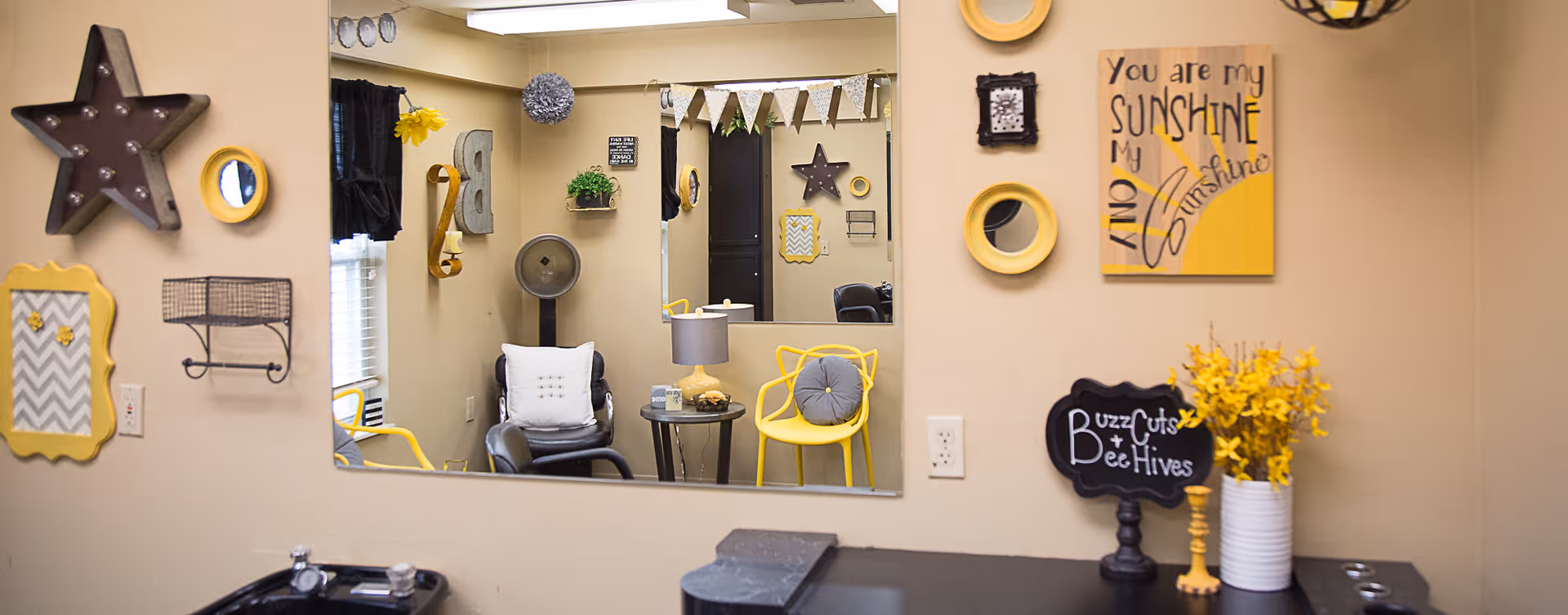 Interior salon-style room with a large wall mirror, yellow and gray decor, seating, and wall signs including "You are my sunshine."