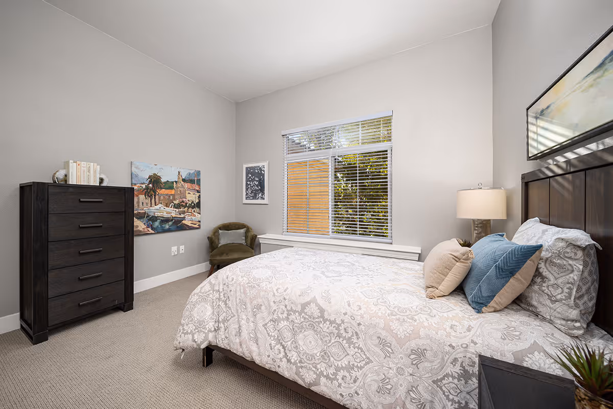 A bedroom with a large bed covered in a patterned white and gray comforter and several pillows. There is a dark wooden dresser with books and decorative items on top, a green upholstered chair, and two framed pictures on the light gray walls. A window with white blinds lets in natural light, and a bedside table with a lamp is next to the bed.