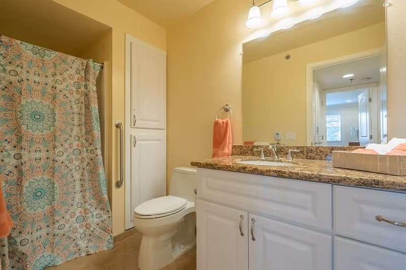A bathroom with a white toilet, a granite countertop with a sink, white cabinets, a large mirror, and a shower with a colorful patterned curtain. There is a coral-colored hand towel hanging on a ring next to the sink, and the walls are painted a warm beige color.