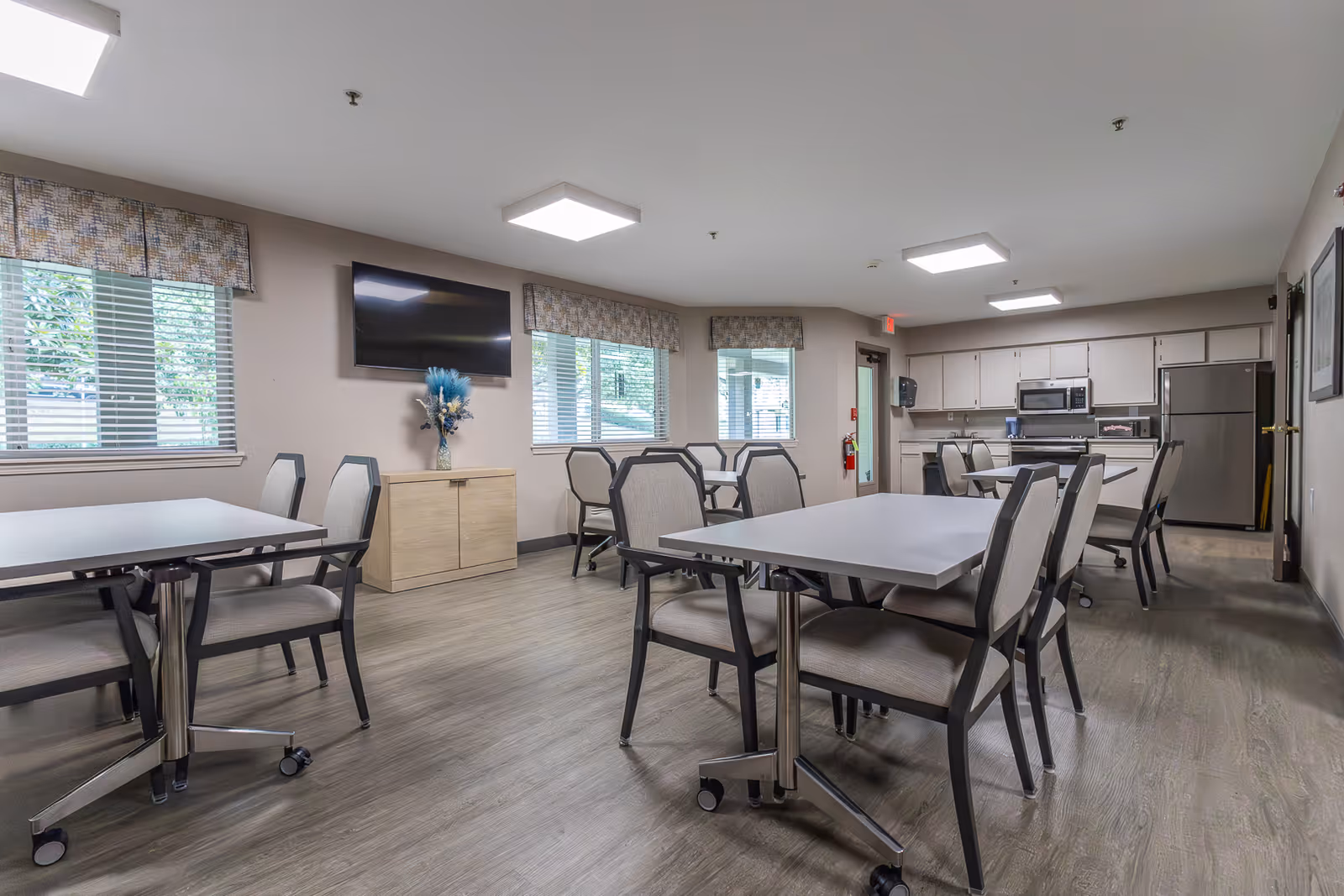 A bright and clean communal dining area with multiple tables and chairs arranged neatly. The room features large windows with blinds and valances, a wall-mounted flat-screen TV, a small cabinet with a decorative vase, and a kitchen area with stainless steel appliances including a refrigerator, microwave, and stove. The flooring is wood-style, and the ceiling has recessed lighting.