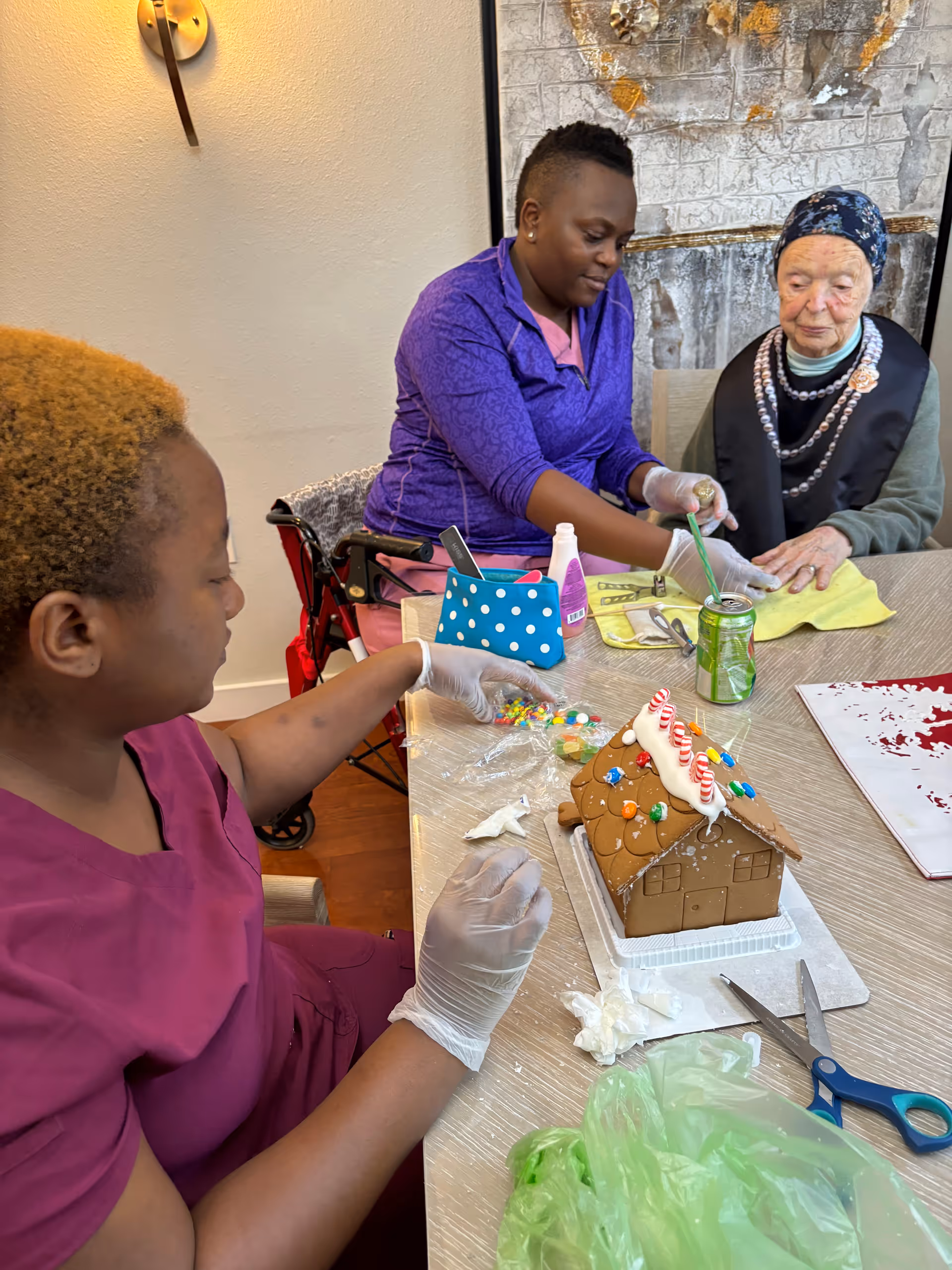Two women sitting at a table engaged in decorating a gingerbread house. One woman wearing a purple top and gloves is placing candy decorations on the gingerbread house, while the other woman, an elderly lady wearing a headscarf and layered necklaces, watches. Various decorating supplies and a can of soda are on the table.