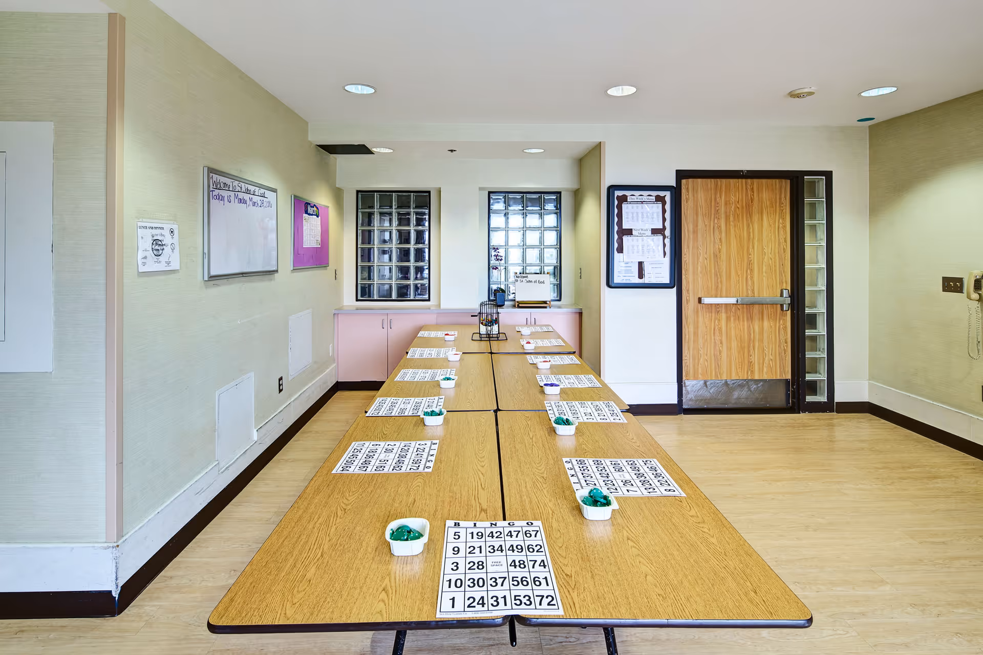 A long table set up for a bingo game with bingo cards and green markers placed in small white containers. The room has light-colored walls, a wooden door, bulletin boards, and glass block windows. The floor is light wood, and the ceiling has recessed lighting.