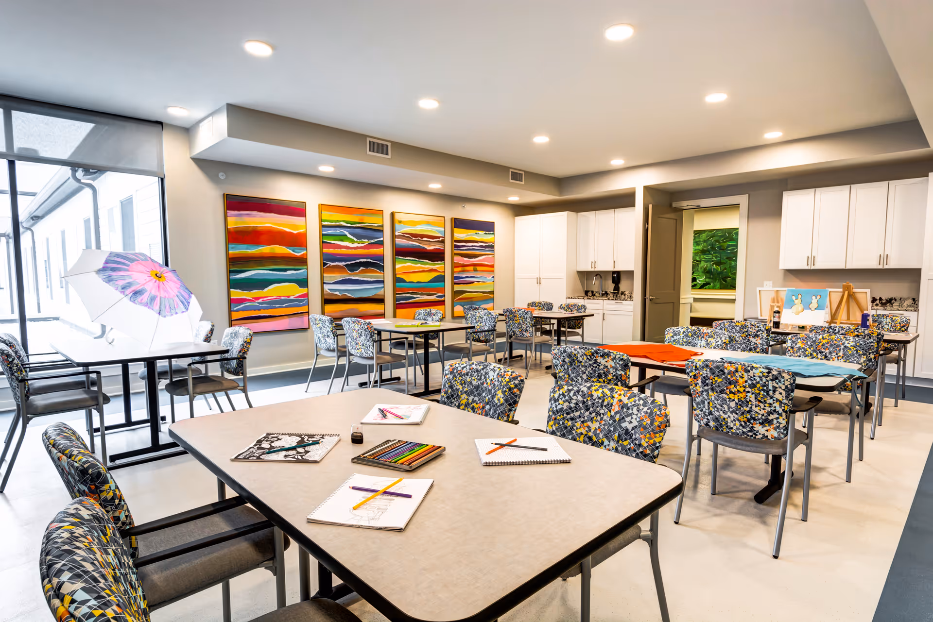 A bright and spacious activity room in a senior living facility with multiple tables and colorful patterned chairs. Art supplies including colored pencils, sketchbooks, and drawings are placed on the tables. The room features modern white cabinetry, vibrant abstract paintings on the wall, and large windows letting in natural light.