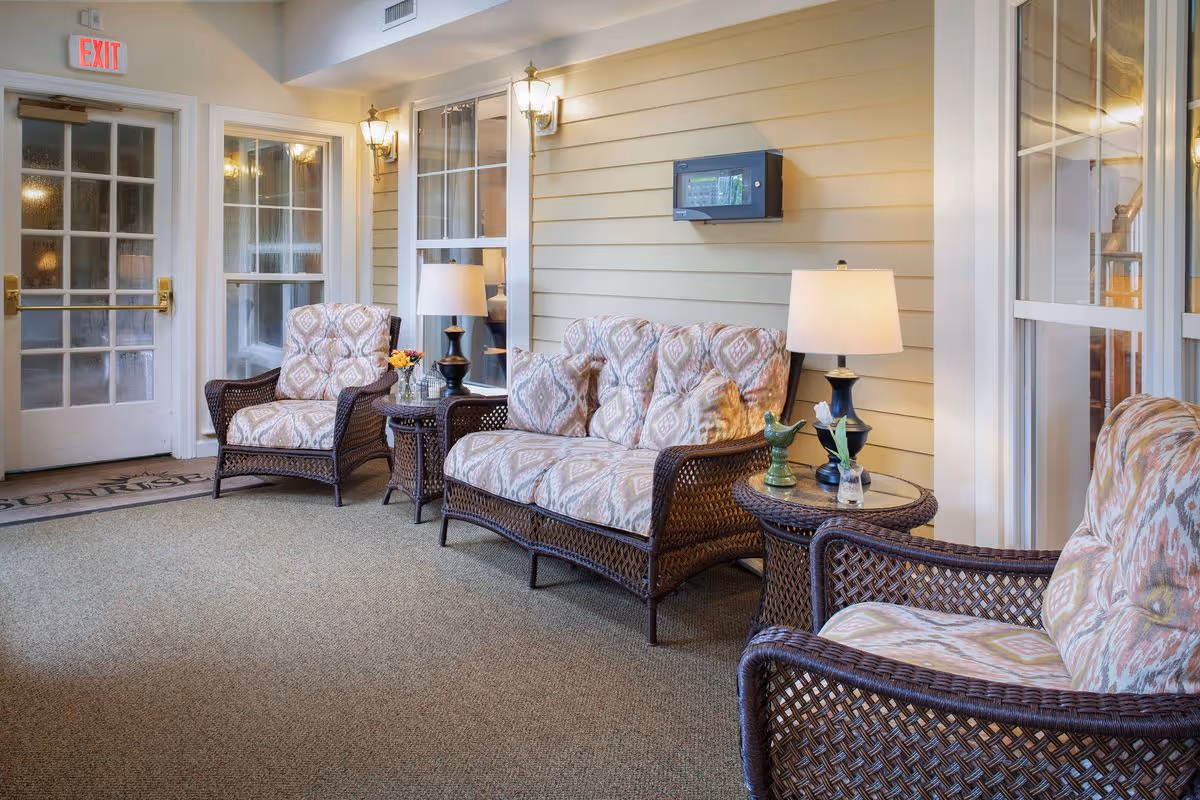 A cozy indoor seating area with wicker furniture including a loveseat and two armchairs with patterned cushions. There are two round wicker side tables with lamps and small decorative items. The walls are light yellow with white trim, and there are windows and a glass door with an exit sign above it.