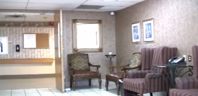 A waiting area inside a facility with several upholstered chairs arranged along the walls. There are two framed pictures on the wall and a small table with a telephone on it. The floor is tiled, and there is a window with a decorative frame on the wall. The background shows a hallway with a bulletin board and a hand sanitizer dispenser.
