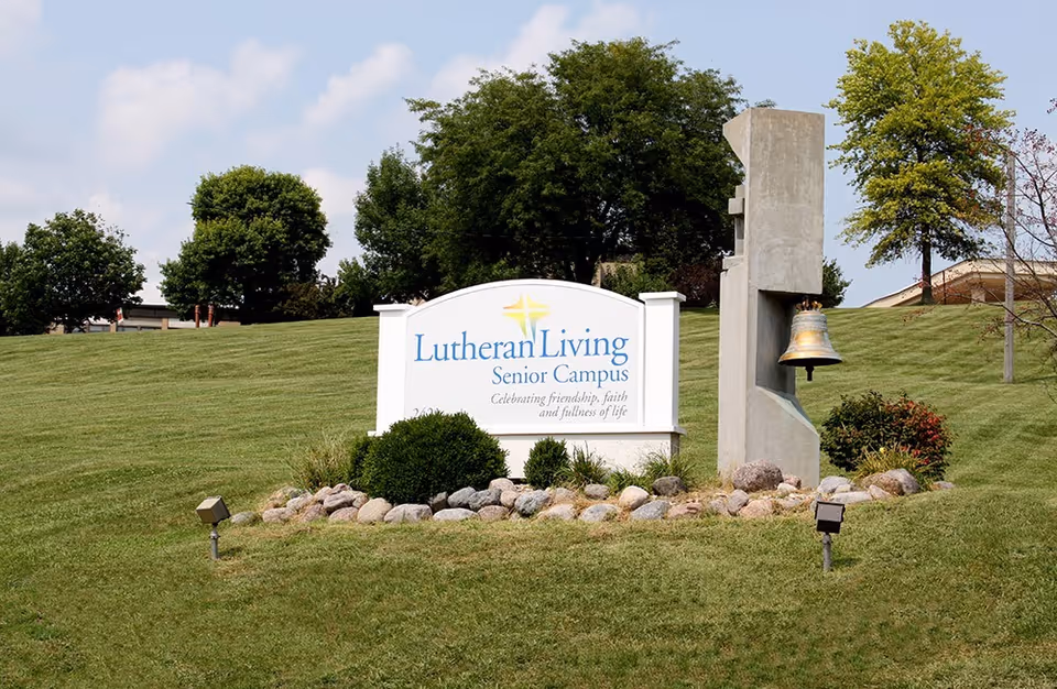 A grassy lawn with a 'Lutheran Living Senior Campus' sign next to a bell sculpture and trees in the background.