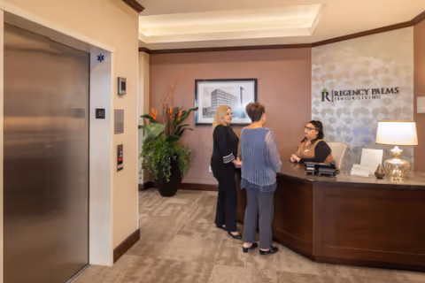 Reception area of Regency Palms Senior Living with a receptionist seated behind a wooden desk talking to two women standing in front of the desk. There is an elevator on the left side and a large plant next to the wall with a framed picture above it. The wall behind the receptionist has the Regency Palms Senior Living logo.