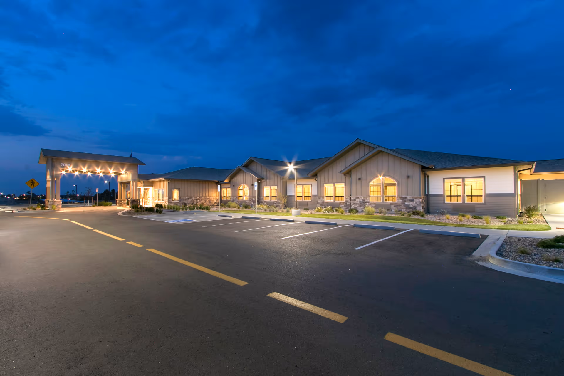Exterior view of a single-story senior living facility building at dusk with lights on inside and a covered entrance with multiple lights. The parking lot in front is mostly empty with marked parking spaces and a clear sky above.