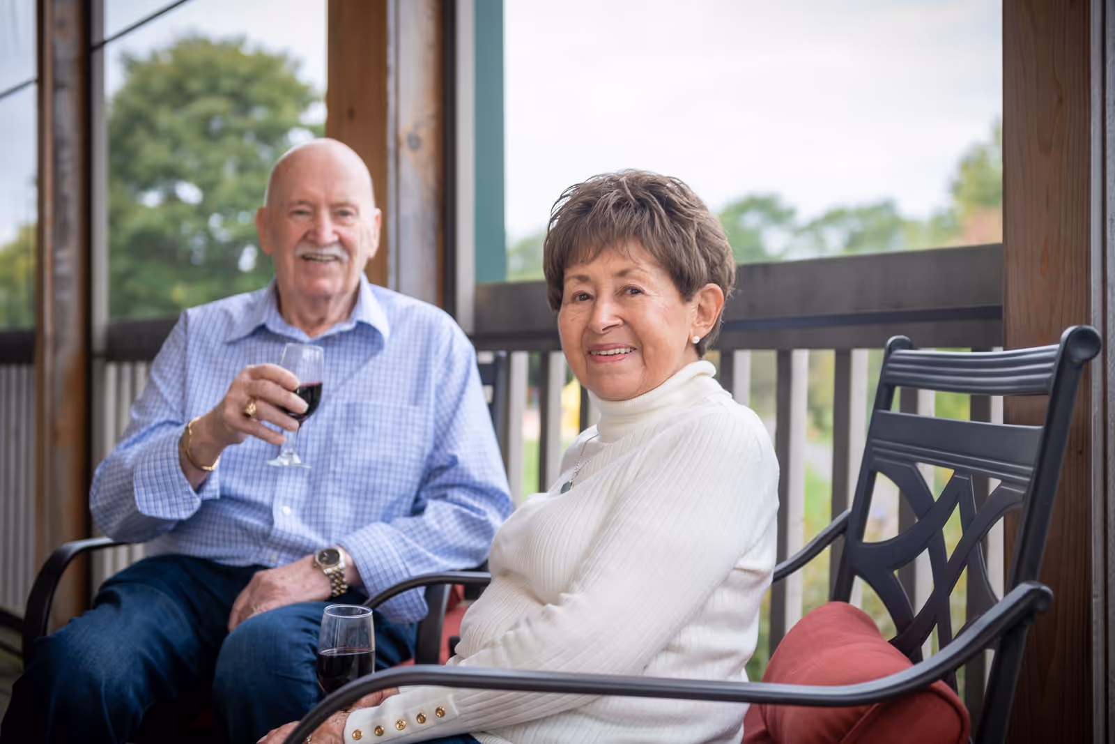 An elderly man and woman sitting on a porch or balcony with wooden railings and greenery in the background. The man is holding a glass of red wine and smiling, while the woman is seated next to him, also smiling and wearing a white sweater.