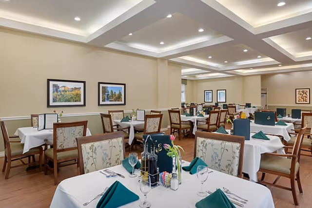 Bright dining room with neatly set tables, white tablecloths, green napkins, and framed artwork on the walls.
