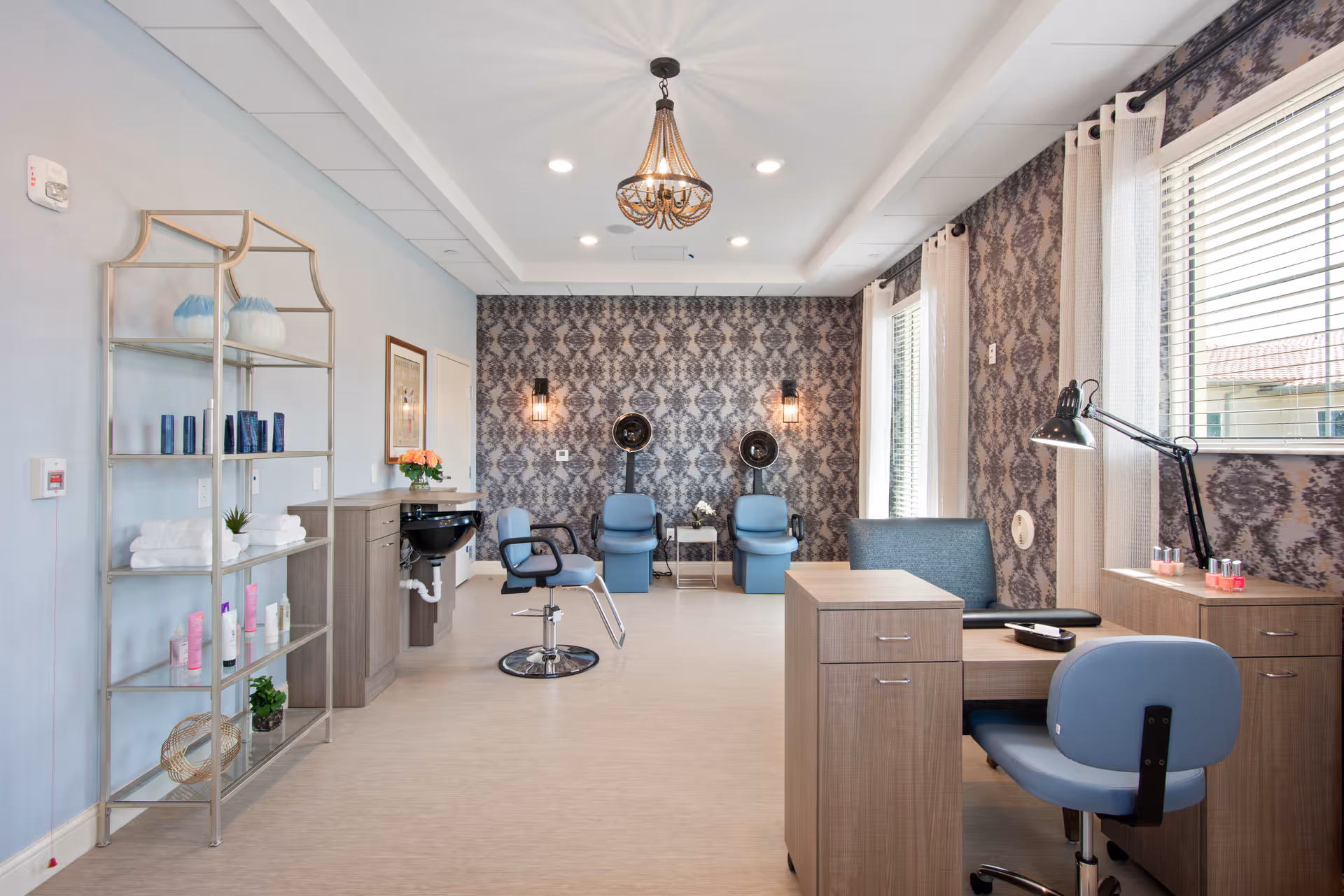 A bright and modern salon room with light blue walls and patterned wallpaper on the far wall. The room features two blue salon chairs with hair dryers mounted on the wall behind them, a styling chair in front of a sink, a shelving unit with towels and hair products, and a manicure station with a blue chair and desk lamp near a window with blinds.