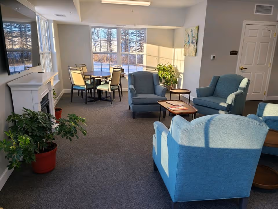 A bright and cozy common area in a senior living facility with blue upholstered armchairs arranged around wooden coffee tables. There is a small dining table with chairs near large windows letting in natural light. Potted plants and a wall-mounted TV above a white fireplace add to the welcoming atmosphere.