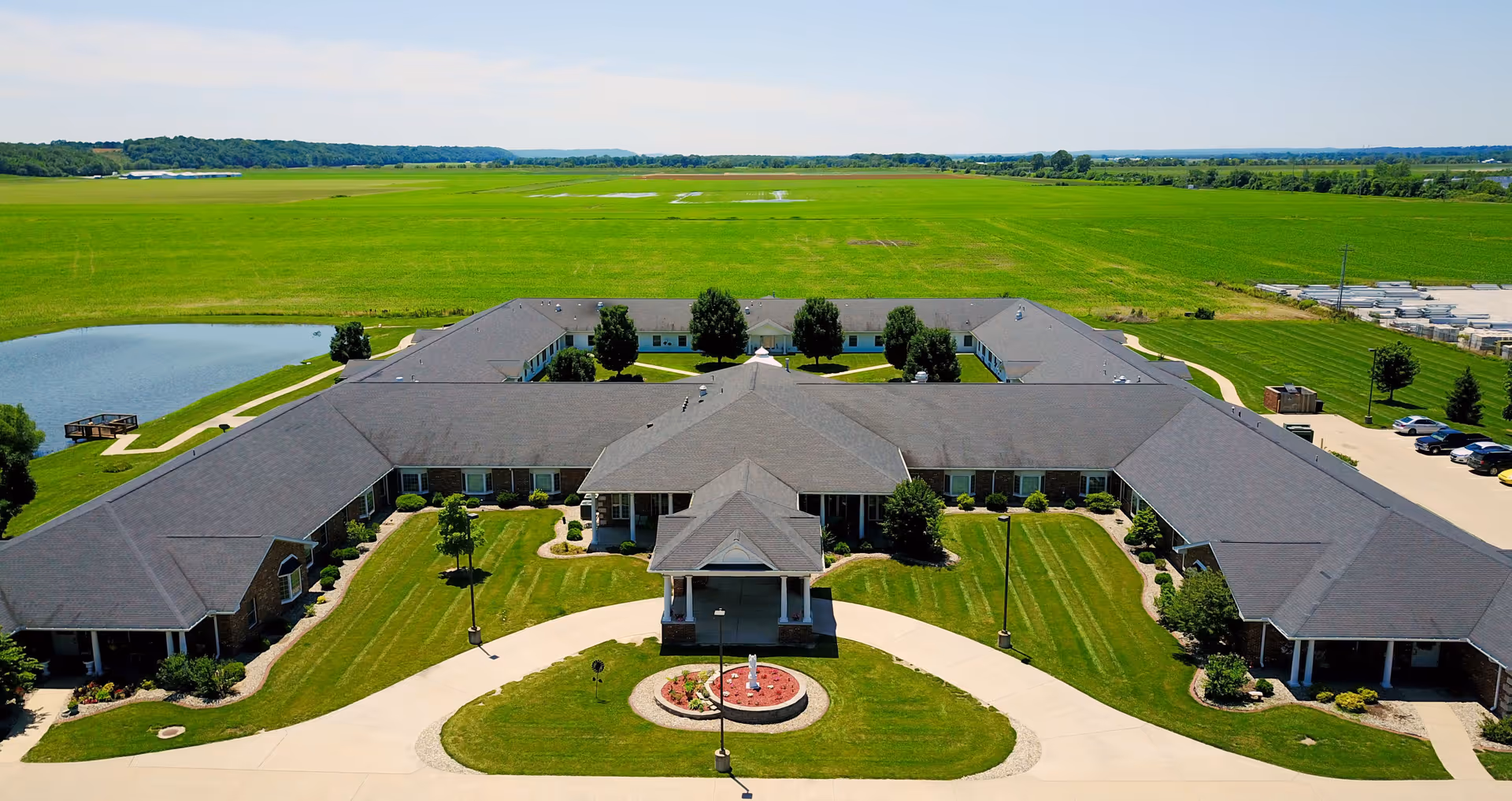 Aerial view of Garden Place Columbia, a large single-story building with a U-shaped layout surrounded by green lawns and trees, with a small pond on the left side and a parking area on the right side under a clear blue sky.