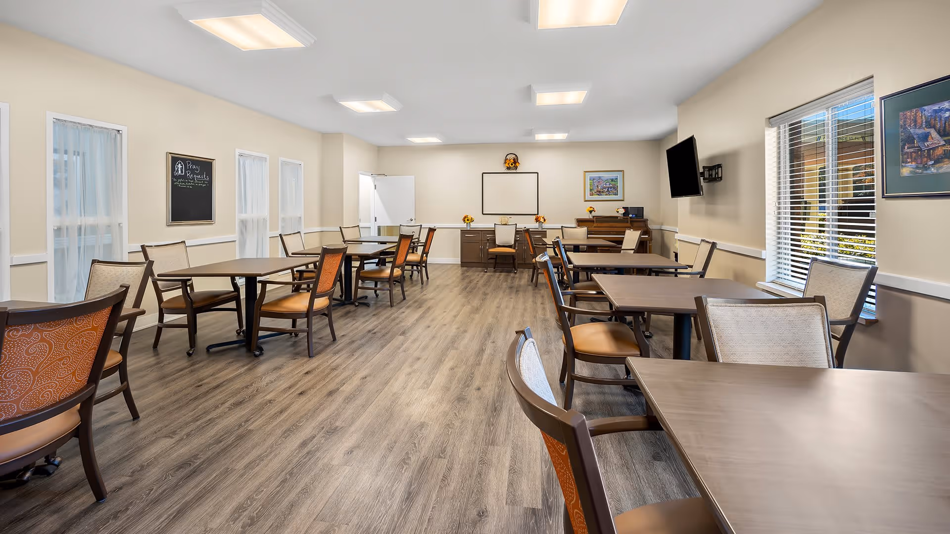 A bright and spacious dining room with multiple tables and chairs arranged neatly. The room has wood-style flooring, beige walls, and several windows allowing natural light to enter. There is a wall-mounted TV on the right side and framed artwork on the walls. At the far end, there is a whiteboard and a cabinet with decorative items.
