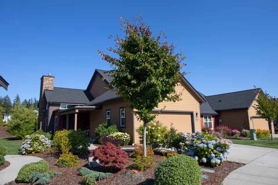 Exterior view of a residential-style building with a garage, surrounded by well-maintained landscaping including various shrubs, flowering plants, and a tree under a clear blue sky.