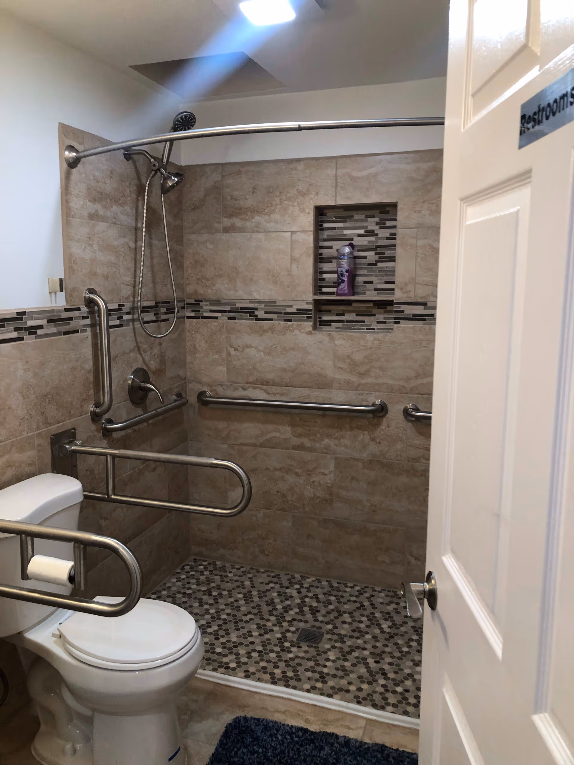 Accessible bathroom with a toilet and a walk-in shower. The shower has a handheld showerhead, grab bars on the walls, and a built-in shelf holding a bottle of shampoo. The toilet has a grab bar with a toilet paper holder attached. The floor has small hexagonal tiles, and there is a blue bath mat in front of the toilet. The door is partially open with a sign that reads 'Restrooms'.