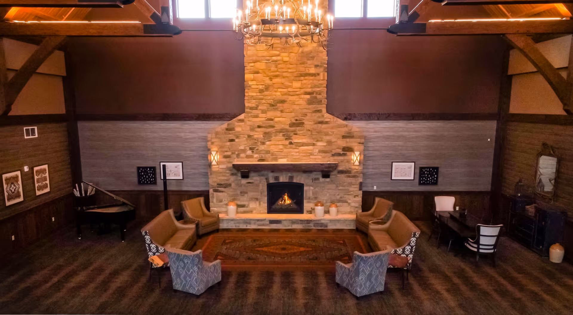 A spacious senior living common area featuring a large stone fireplace with a fire burning, surrounded by comfortable seating including armchairs and sofas arranged in a semi-circle. The room has wooden beams, a chandelier hanging from the ceiling, a piano in the corner, framed artwork on the walls, and a desk with chairs on the right side.