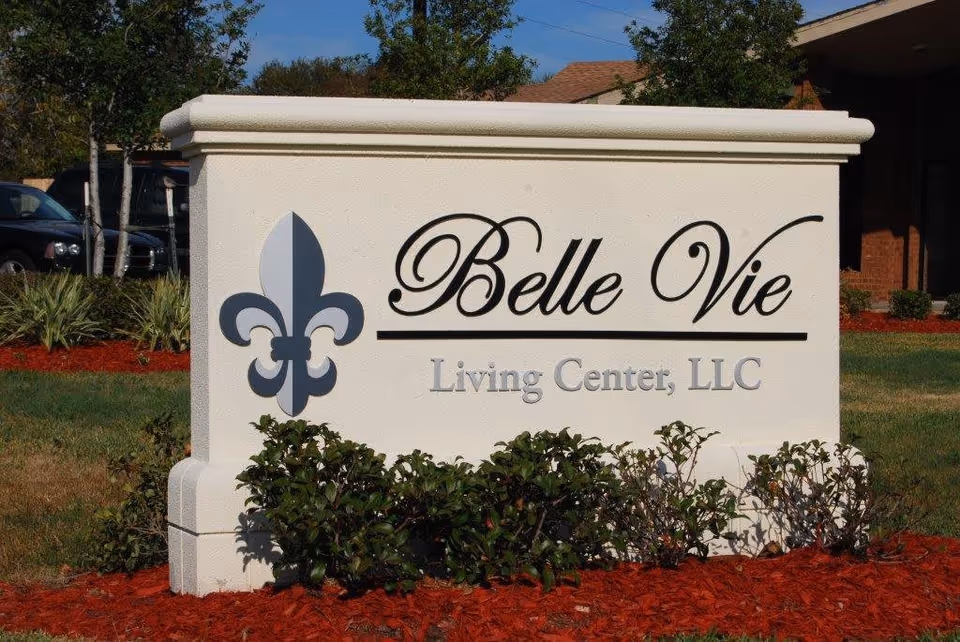 Outdoor sign for Belle Vie Living Center, LLC with a fleur-de-lis symbol on the left side, surrounded by landscaping with red mulch and green bushes.