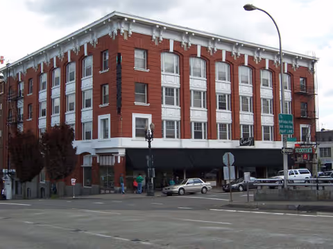 A large, four-story red brick building with white trim and multiple windows on each floor. The building is located at a street corner with several cars parked nearby and a few people walking on the sidewalk. There are street lamps and traffic signs visible around the building.