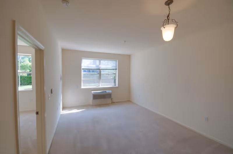 Empty room with beige carpet and cream-colored walls, a window with a white blind partially open, an air conditioning unit below the window, and a hanging light fixture on the ceiling. A doorway on the left side leads to another room with a window showing greenery outside.