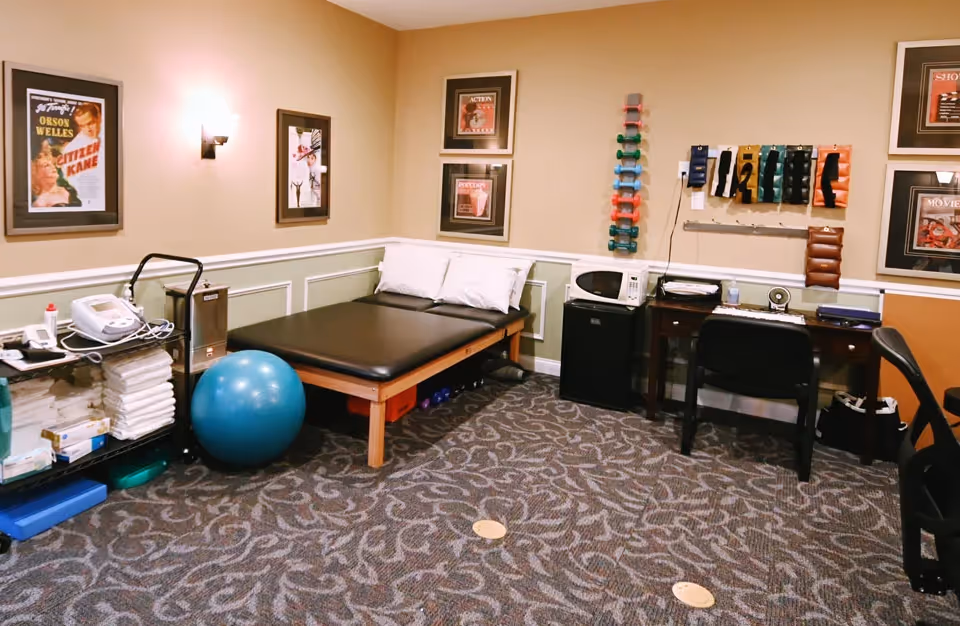Therapy room with a treatment table, exercise ball, bands, and a desk against beige walls.