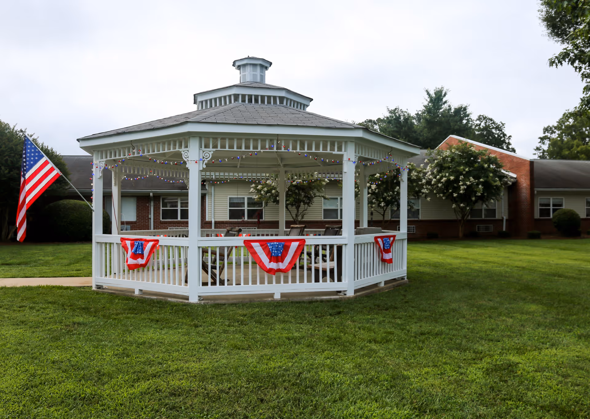 A white gazebo decorated with red, white, and blue bunting and string lights, situated on a green lawn in front of a single-story building with trees and an American flag nearby.