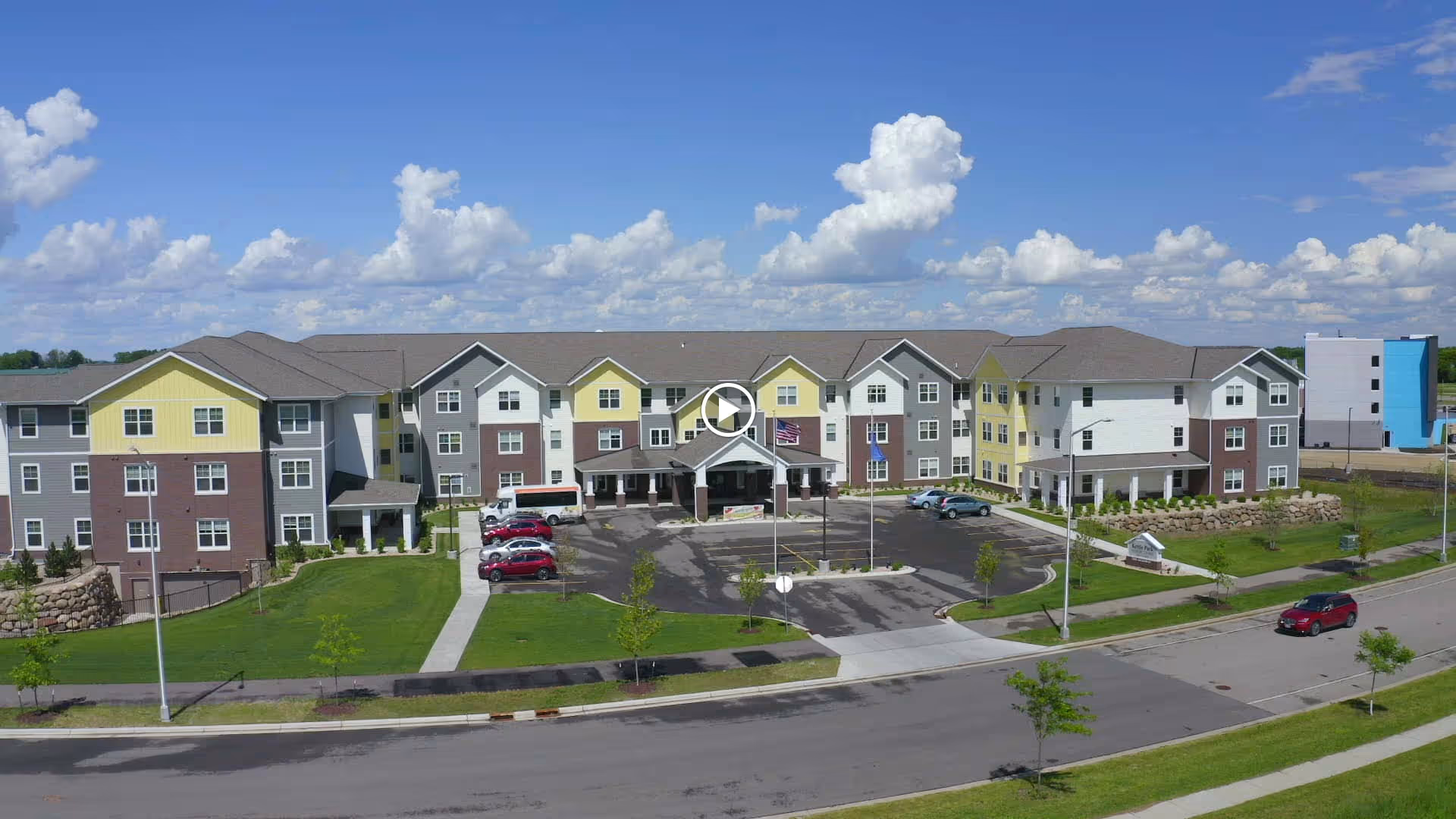 Three-story senior living building with a central entrance, parking lot, small lawn and flagpoles under a blue sky.