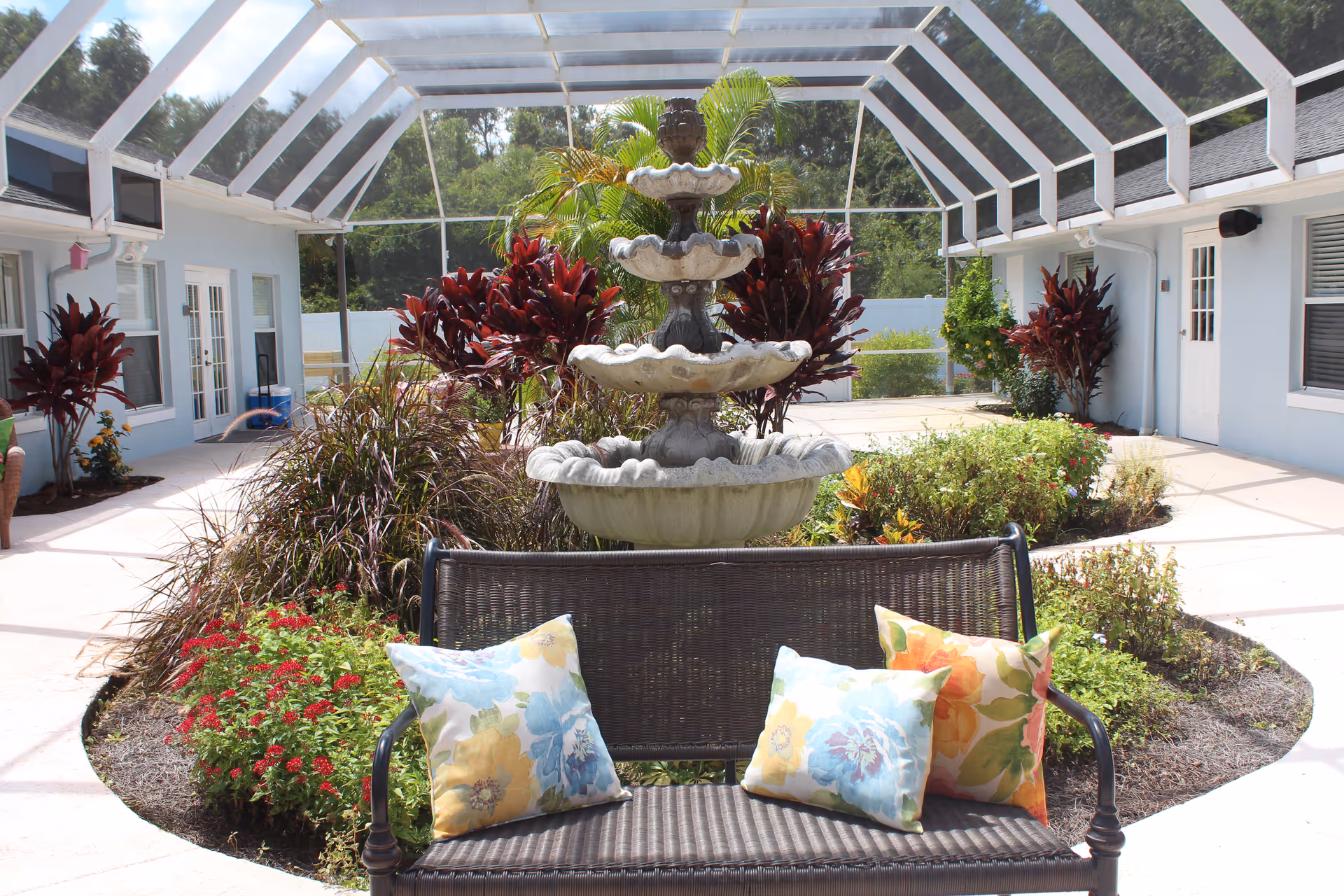 Outdoor courtyard area with a multi-tiered stone fountain surrounded by plants and flowers. A brown metal bench with three colorful floral cushions is in the foreground. The courtyard is enclosed by a screened roof and light blue buildings with windows and doors.