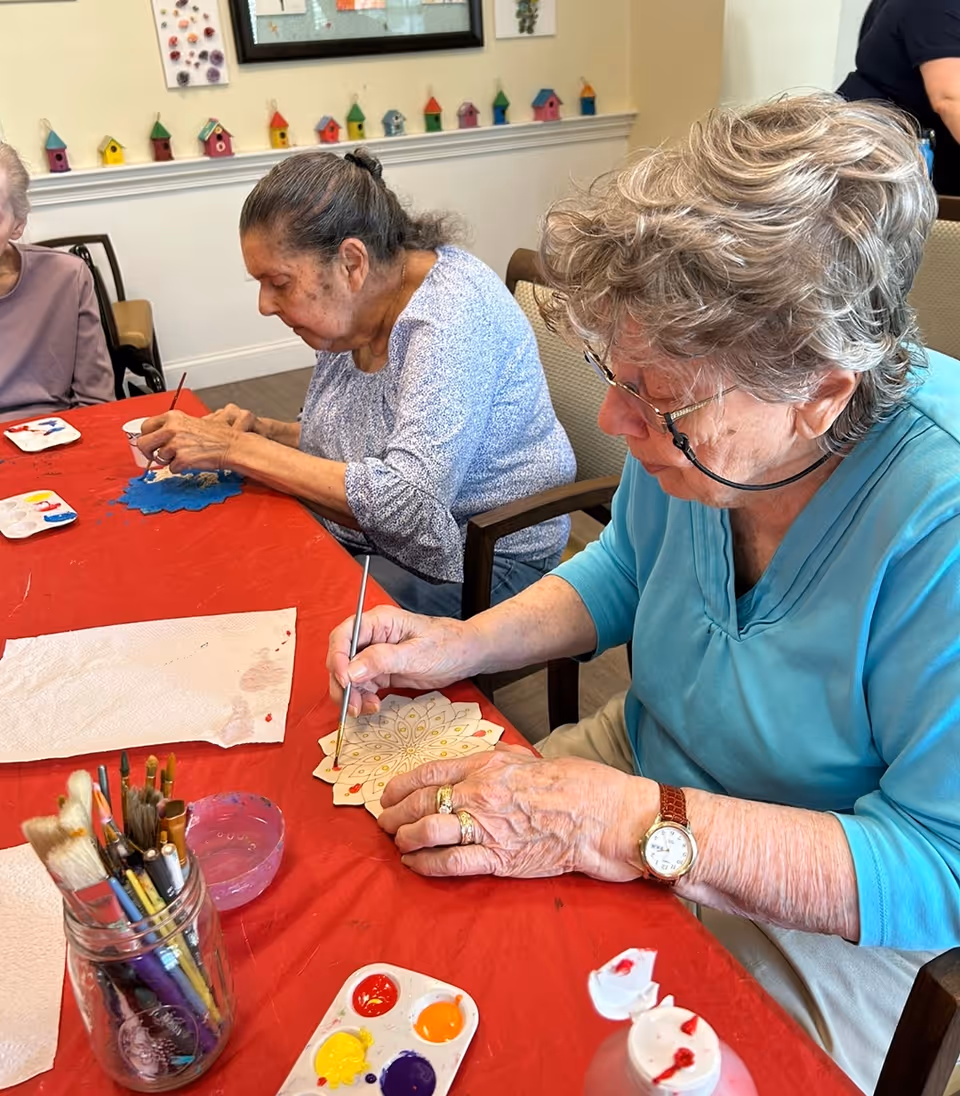 Two elderly women sitting at a table covered with a red tablecloth, painting decorative wooden pieces with small paintbrushes. Various paint colors and brushes are visible on the table. The room has light-colored walls with small colorful birdhouse decorations on a shelf in the background.