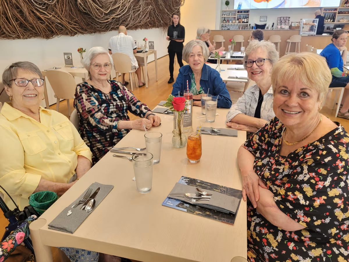 Five elderly women sitting around a dining table in a bright, modern dining room. The table is set with glasses of water, utensils wrapped in napkins, and a vase with a red rose. In the background, other people are seated at tables and a server is standing. The room has light-colored walls and wooden flooring.