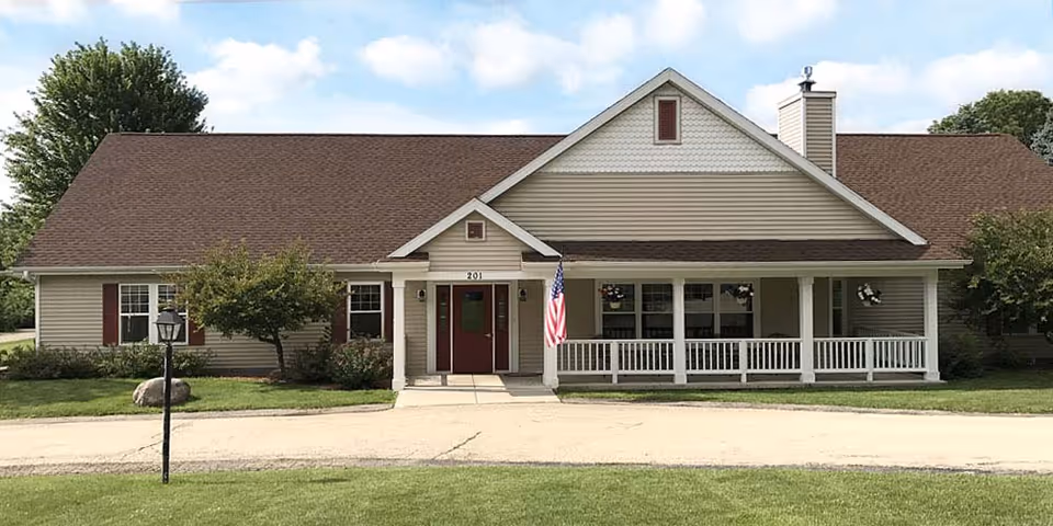 Single-story building with beige siding and a brown roof, featuring a covered porch with white railings and an American flag near the entrance. The building is surrounded by green grass, small trees, and shrubs under a partly cloudy sky.