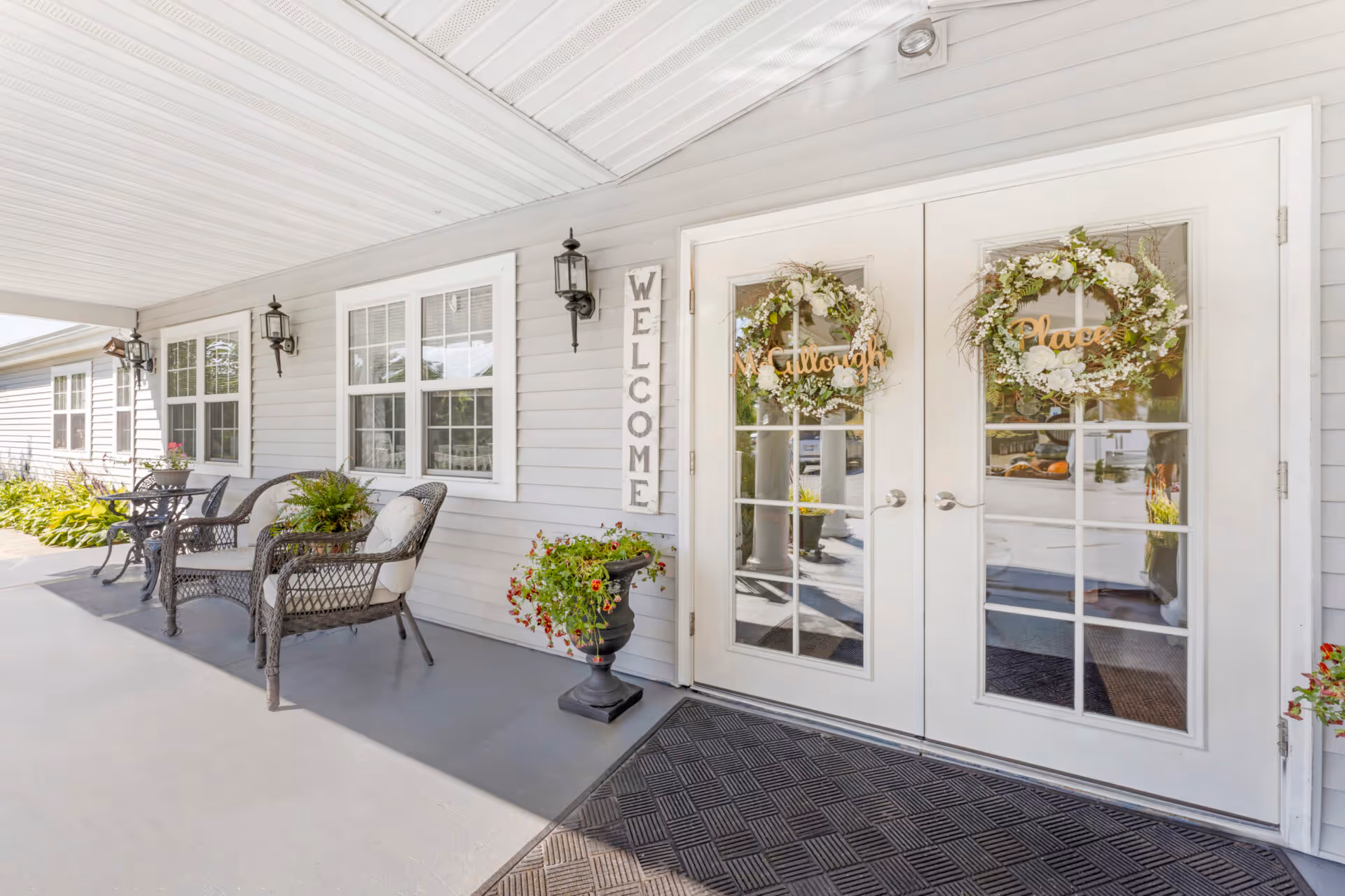 Covered front porch with wicker seating, potted plants, a vertical "WELCOME" sign and double glass doors adorned with wreaths.