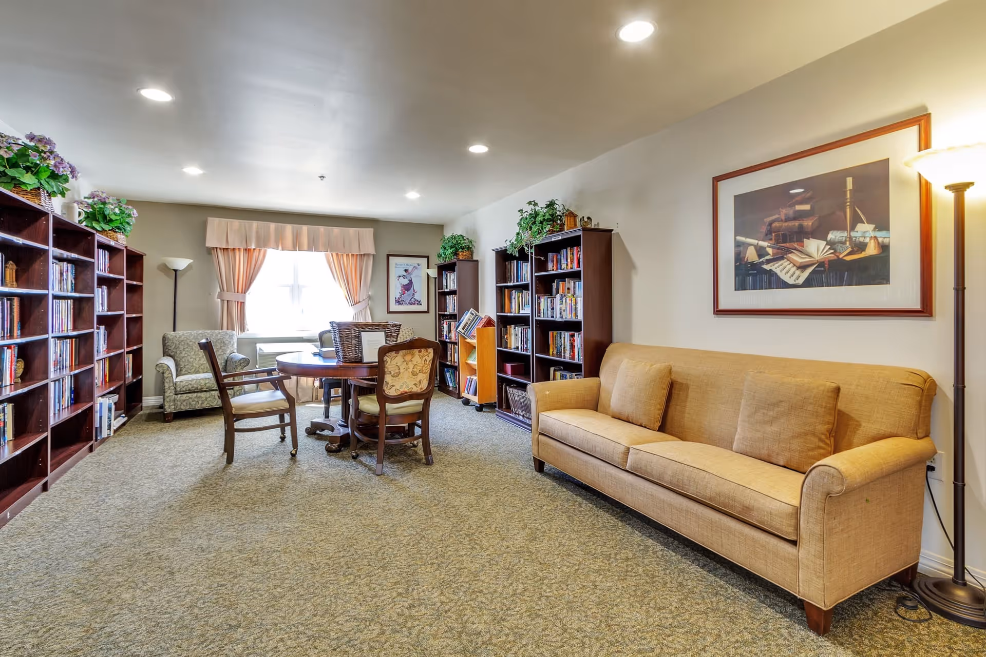 A cozy common room with a beige sofa, a round wooden table surrounded by chairs, bookshelves filled with books, a green armchair near a window with peach-colored curtains, and decorative plants on top of the shelves. The room is well-lit with ceiling lights and a floor lamp.