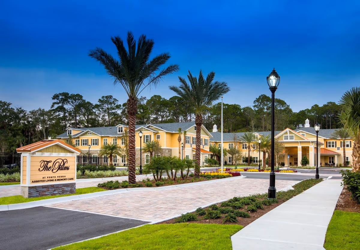 Exterior view of a yellow senior living campus with palm trees and a sign.