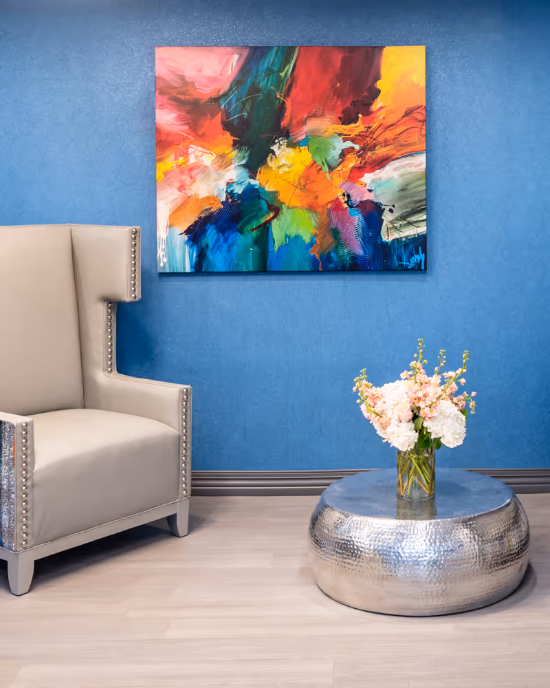 A modern interior corner featuring a beige armchair with nailhead trim, a round hammered metal coffee table with a glass vase of pink and white flowers, and a colorful abstract painting on a blue textured wall.