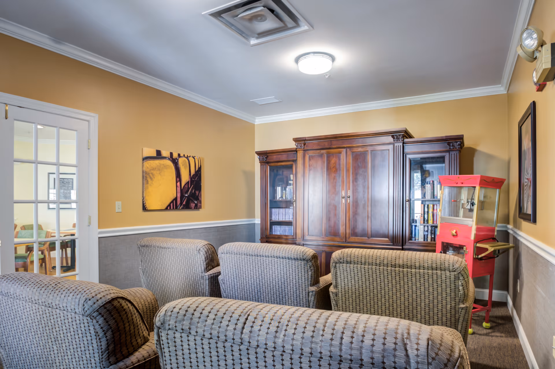A cozy living room area with four patterned armchairs arranged in two rows facing a large wooden entertainment cabinet. To the right of the cabinet is a red popcorn machine. The walls are painted yellow with a gray wainscoting, and there is a framed picture on the right wall. A glass door on the left leads to another room with tables and chairs.