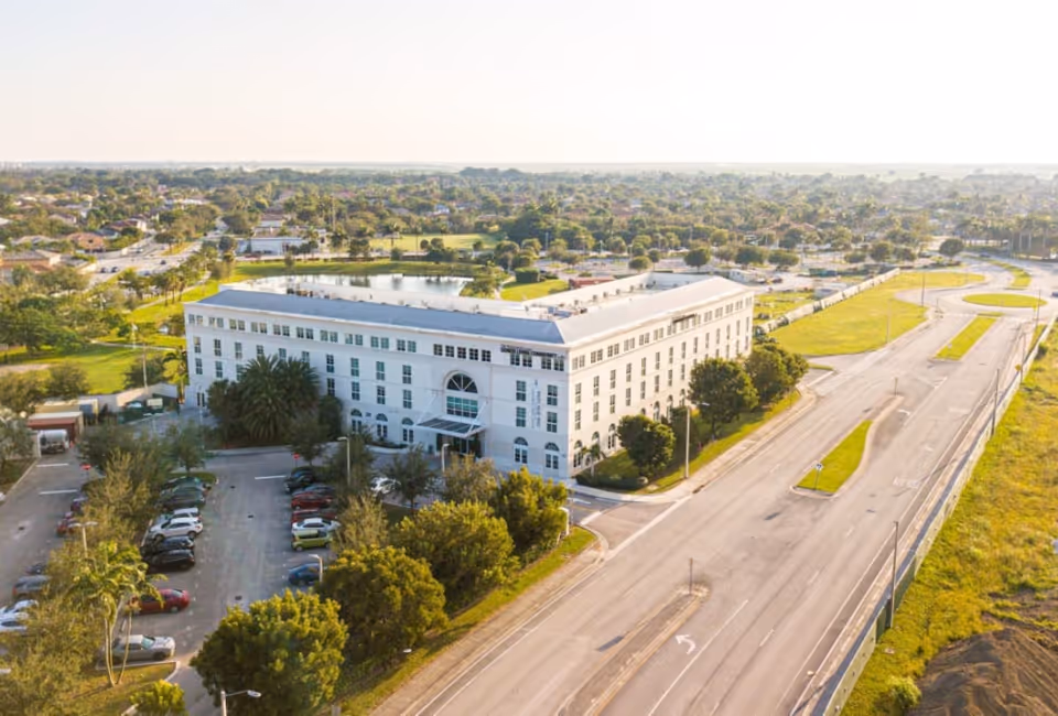 Aerial view of a large white multi-story building surrounded by trees and parking lots, with a pond and residential neighborhood in the background under a clear sky.